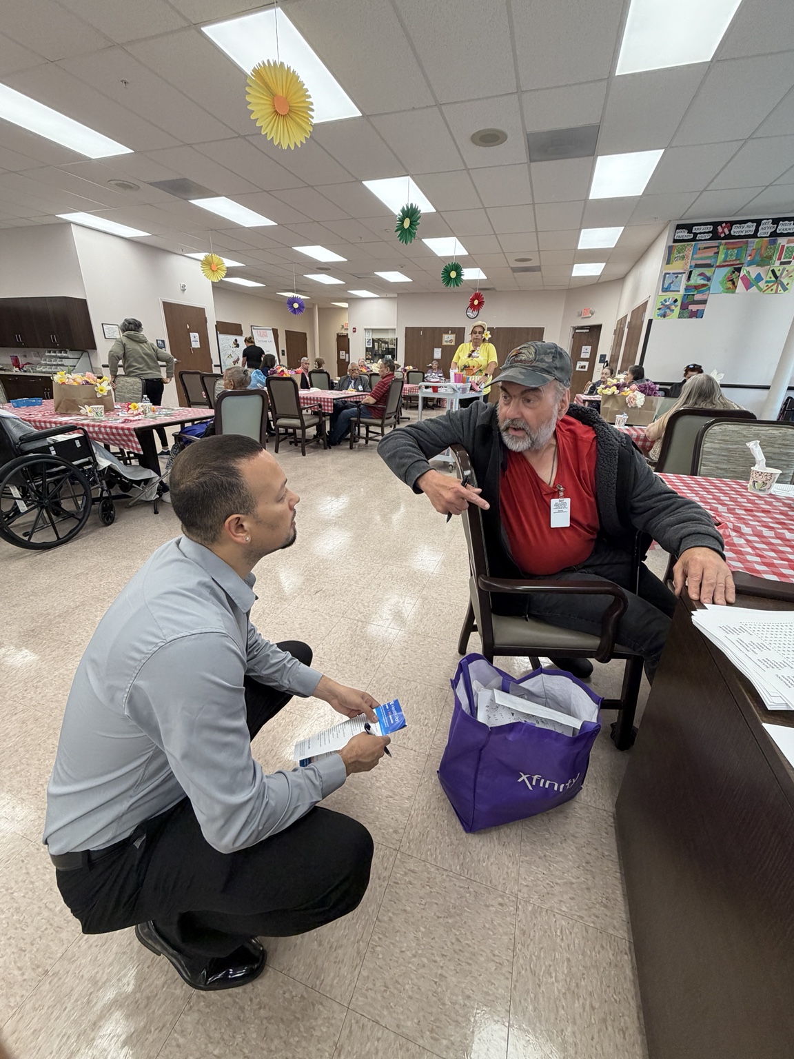 Jaime crouching beside a seated older neighbor during a conversation at Senior LIFE