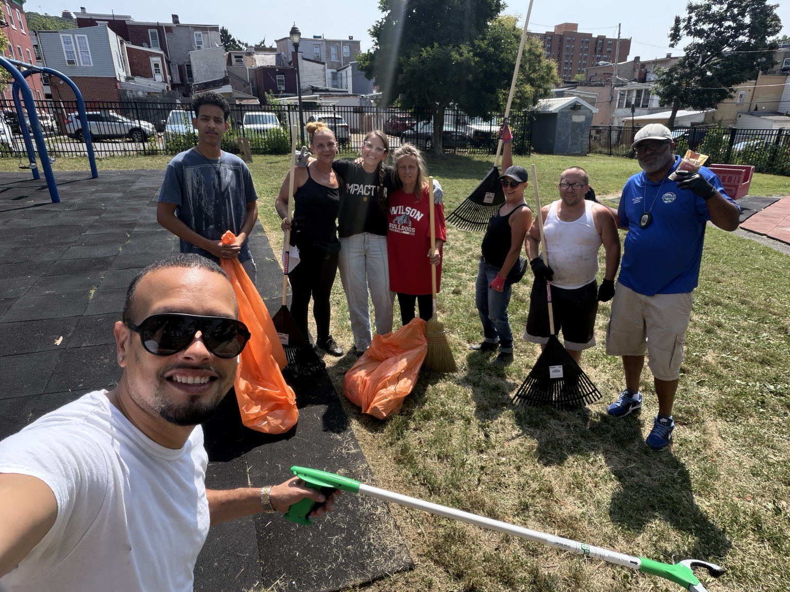 Jaime taking a group selfie with volunteers holding rakes, litter pickers, and trash bags at a playground cleanup